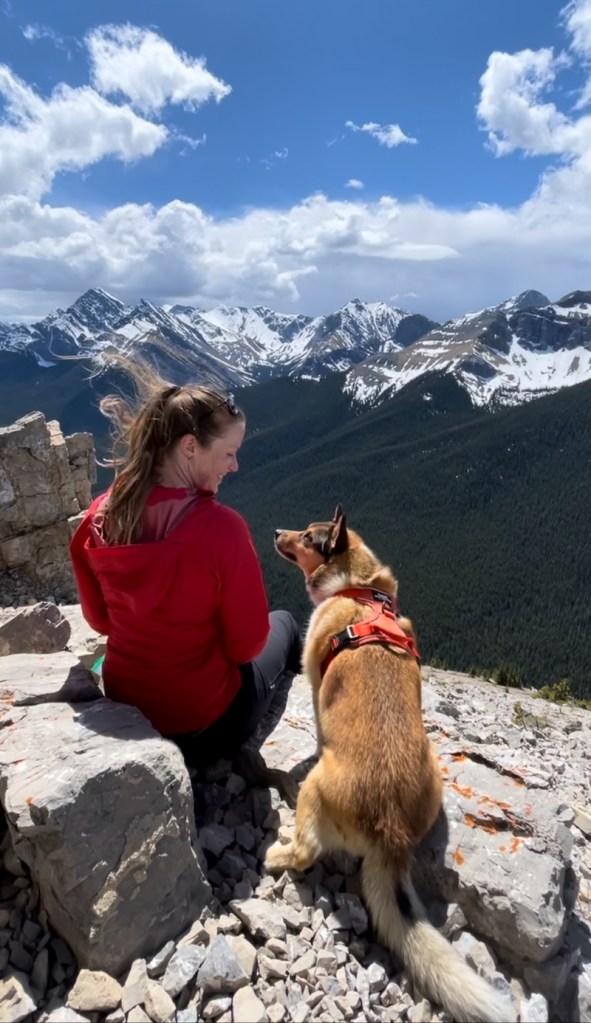 Woman in red jacket smiling at brown dog; both are sitting on top of cliff with mountains in view.